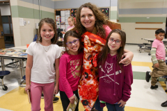 Three children posing with a woman in a pizza costume in a classroom setting.
