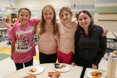 Three young girls posing in front of a table with pizza, smiling at the camera.
