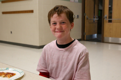 Young boy in pink shirt sitting at table in school cafeteria.