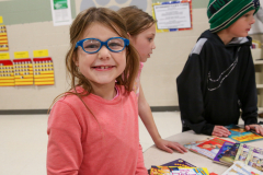 Girl in glasses smiling at camera, standing next to table with books.