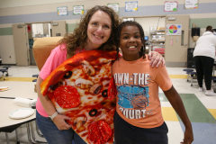Woman holding a pizza in front of her, posing for a picture with a smiling boy behind her.
