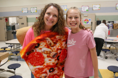 Woman posing with cardboard pizza cutout in school cafeteria.