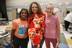 Three people posing in front of a pizza shaped like a slice of pizza. The person on the left is wearing a shirt that says