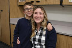 Woman in plaid shirt and glasses smiling at camera next to a young boy wearing glasses and a blue top. They are standing in