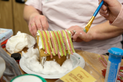 Gingerbread house decorated with candy, being crafted by a person using blue paintbrush.