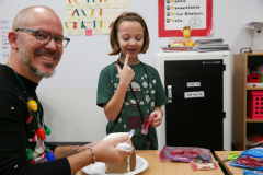 A man in a festive sweater is making gingerbread houses with a child at a table.