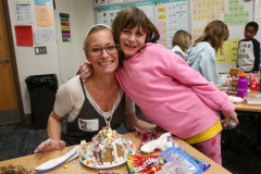 Woman wearing glasses hugging child at gingerbread house table with candy decorations.