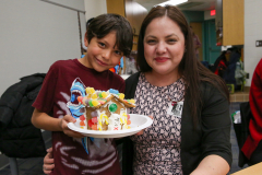 Woman holding a plate of gingerbread houses. A boy standing next to her. They are both smiling and posing for the picture.
