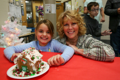 Two people posing in front of a gingerbread house at a Christmas event.