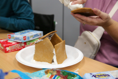 Person holding a gingerbread house in front of a table with various snacks and drinks.