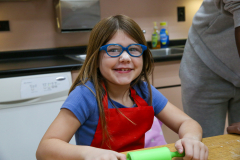 Girl in glasses sitting at kitchen counter, smiling, wearing blue shirt.