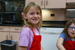 Girl in kitchen wearing apron, smiling at camera.