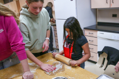 People in a kitchen preparing food. A woman is rolling dough on a countertop while a child watches. The kitchen has a white