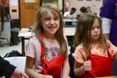 Three children in a classroom setting, two girls wearing aprons and one boy, all smiling at the camera.