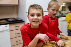 Two young boys in kitchen, one smiling at camera, both wearing aprons, making cookies.