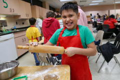 Boy in red apron holding rolling pin with flour on it.