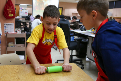 Two children in a classroom setting, one boy wearing an apron working on a project at a table while another child looks on.