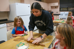 Woman in kitchen, making gingerbread cookies, surrounded by children watching her work.