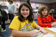 Two young girls sitting at a table in a classroom setting, engaged in an activity. One girl is wearing a yellow shirt and
