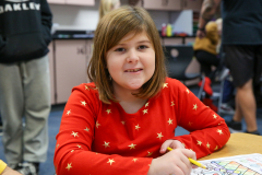 Girl in red shirt sitting at a table, writing on paper, with a smile on her face.