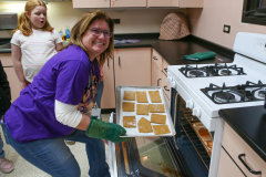 Woman in purple shirt leaning over tray of gingerbread cookies on stove top oven.