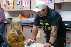 Man in black shirt and white hat standing next to a table with a child. The man is holding a small object in his hand.
