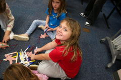 Children sitting on floor surrounded by craft supplies, smiling at camera.