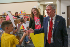 Man in suit shaking hands with children. They are in front of a school building. He is smiling and appears to be greeting