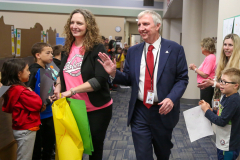 Two people walking in a hallway, waving to children who are watching them. A man wearing a suit and tie is shaking hands