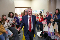 Man in suit standing amidst a group of people clapping, smiling, and holding signs; he appears to be addressing an audience