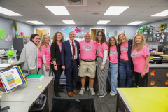 Group of people standing in an office, posing for a picture, all wearing pink shirts with a logo on them.