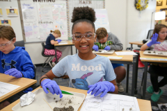 Girl in blue shirt smiling at camera, wearing glasses and gloves, standing in front of table with owl pellet dissection