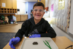Boy in goggles at a table wearing gloves and looking at an owl pellet.