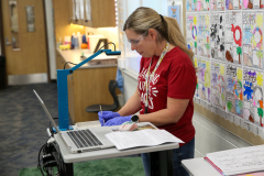 Woman in red shirt standing at desk, signing paperwork, wearing black gloves.