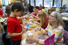An elderly woman is sitting at a table, surrounded by children who are participating in a gingerbread house decorating