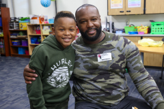 Man wearing camouflage shirt posing with young boy in a classroom setting.