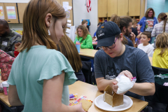 Two people at a table, one building gingerbread houses while the other watches.