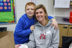 Woman in a grey hoodie sitting at a table with a young boy on her lap. They are both smiling and looking towards the camera.