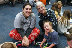 Three people sitting on floor in classroom setting. Woman wearing red pants and black shirt smiling at camera. Child wearing