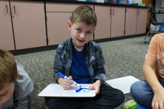 Young boy sitting at a table in a classroom, writing on a whiteboard with a blue marker, surrounded by other children who
