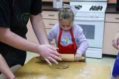 Small child in red apron at kitchen counter, making gingerbread cookies with adults.