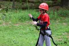 Girl in red helmet holding onto rope, standing on grass, wearing harness.