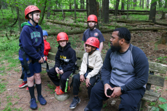 People sitting on a bench in a forested area wearing helmets and holding helmets. A man standing behind them is smiling and