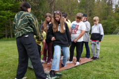 Group of people standing around a wooden board on the grass, possibly participating in an outdoor activity or event.