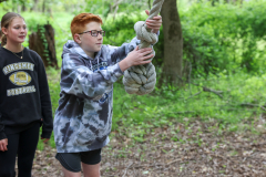 Three people in a wooded area, one person holding a snake wrapped around a stick.