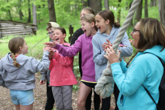 Children at a camp, interacting with an adult, in a group setting, outdoors.