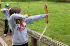 Girl in a white hoodie holding a bow and arrow, practicing archery at camp.