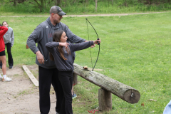 Two people at a shooting range, one holding a bow, the other teaching.