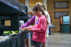 Two girls standing at a buffet table in a cafeteria, preparing food.