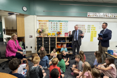 Two adults and a group of children in an educational setting, one adult is gesturing while speaking to the students.